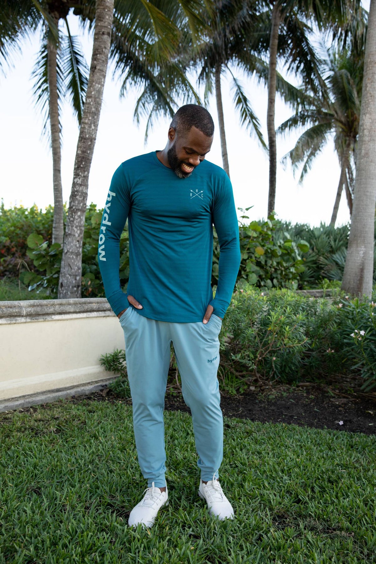 Man posing in dark blue long sleeve breathable shirt with hydrow on right sleeve and crossed oar logo on upper left chest hangs in pockets of teal pants