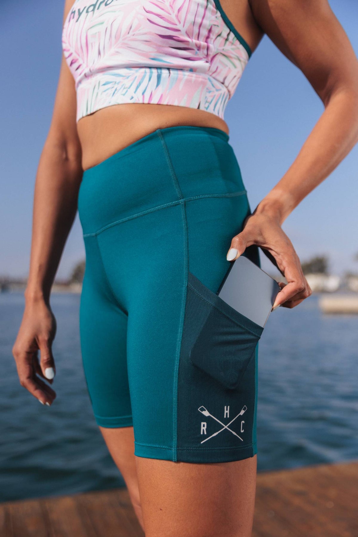 photo of female model posing on boat dock wearing lake lucerne bike shorts and miami tropic racerback bra. showcasing pocket on left leg with cellphone being inserted. rhc crossed oar logo on pocket in white. 