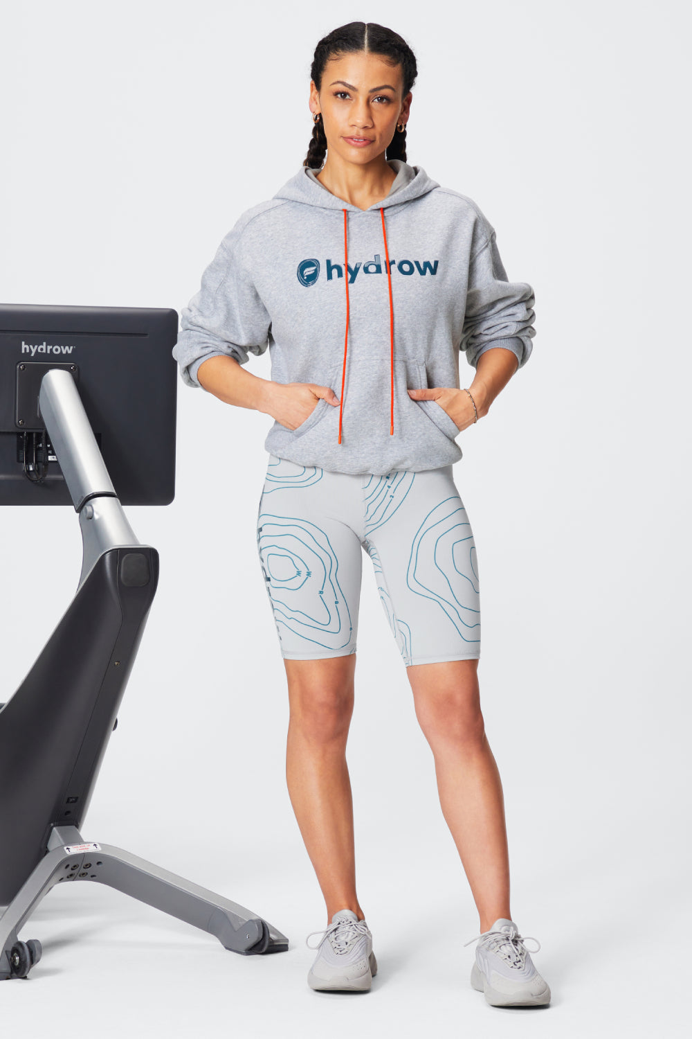 woman standing next to hydrow rower wearing the forever fleece hoodie and the powerhold short